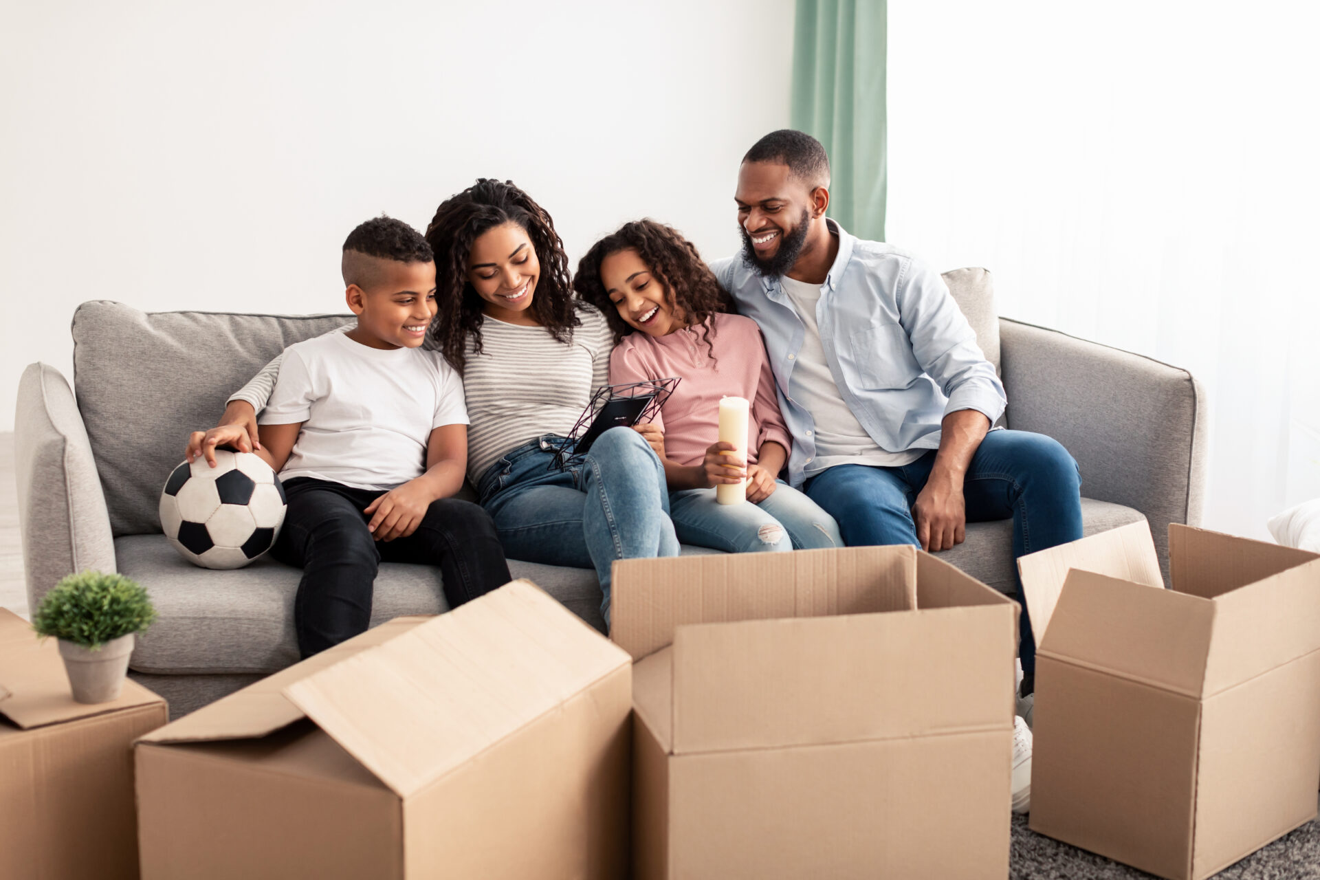 Portrait of smiling black family of four people sitting on the sofa in living room, moving out or relocating, unpacking cardboard boxes, holding picture frame and looking at their photo together